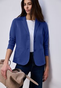 Femme portant un blazer bleu sur une chemise blanche et un jean foncé, tenant un grand sac en daim beige avec une bandoulière.