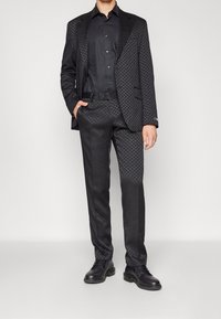 Černý vzorovaný oblek z texturované tkaniny, který obsahuje dobře střiženou bundu a kalhoty v odpovídajícím stylu, doplněn�ý černou košilí na knoflíky a obuví.