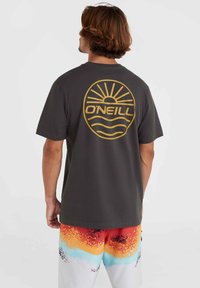 Camiseta gris oscuro con un gran logo de sol dorado y ola en la parte posterior, combinada con shorts de degradado colorido que presentan patrones en rojo, naranja y azul.