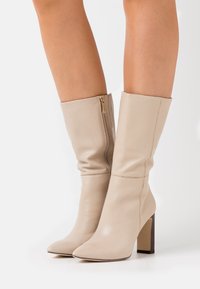 Beiga läderknäboots med spetsig tå, klack med blockform och detaljerad sidogges. Slät yta med sömdekorationer.