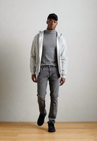 Felpa grigia sportiva sopra un dolcevita grigio scuro, abbinata a jeans slim grigi e sneakers nere. Presenta un cappellino nero e linee pulite.