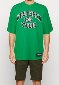 Groene oversized t-shirt met zwarte tekst "WRSTBHVR 030 STUDIO." Korte mouwen, ronde halslijn en een zijlabel met merkinformatie.