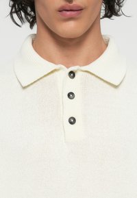 Weißes Strickpoloshirt mit strukturiertem Stoff, umgeschlagenem Kragen und drei schwarzen Knöpfen vorne.