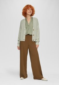 Hellgrauer Strickcardigan mit braunen Knöpfen über einer grünen Bluse und braunen weiten Hosen. Weiße flache Schuhe runden das Outfit ab.