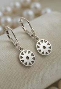 Silverörhängen med cirkulära, utskurna mönster, med ett centralt hål och en låsning med hävarm, visade mot en texturerad tygbakgrund.