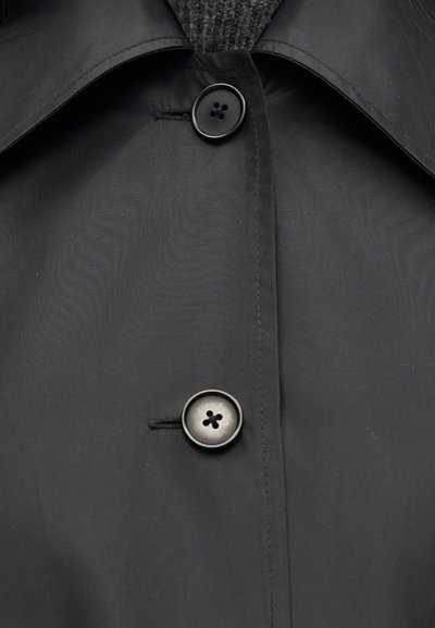 Κοντινό πλάνο στο εμπρός μέρος ενός σκούρου παλτού με δύο μαύρα κουμπιά κλειστά, που δείχνει λεπτομέρειες ραφής και μέρος από μία υφή γκρι εσωτερική στρώση.