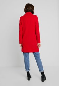 Frau, die mit dem Rücken zur Kamera steht, trägt einen leuchtend roten knielangen Mantel, gekrempelte blaue Jeans und schwarze, Absatz-Ankle-Boots vor einem schlichten grauen Hintergrund.