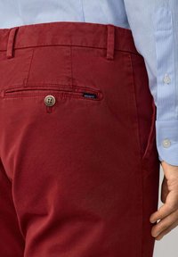 Pantalones de algodón rojos con una textura suave, que cuentan con un bolsillo trasero con cierre de botón y una etiqueta de la marca. Puño de camisa a rayas azules visible.