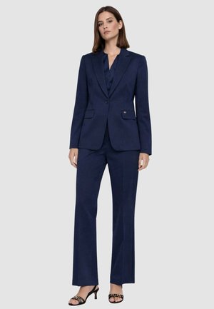 Donna che indossa un completo su misura blu navy con blazer monopetto, pantaloni abbinati, sandali neri con tacco, in piedi davanti a uno sfondo neutro.