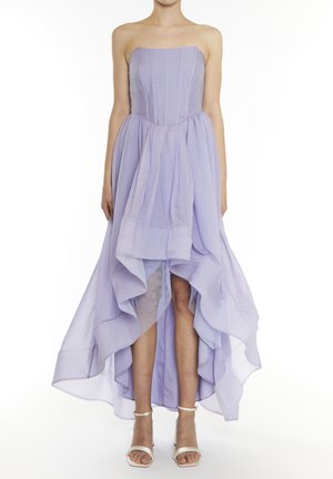 Robe lavande bustier à ourlet asymétrique avec jupe fluide à couches, portée par une personne debout, accompagnée de talons blancs à bride autour de la cheville, sur fond blanc.