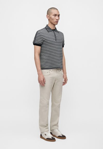 Homme portant un polo gris rayé à manches courtes, un pantalon beige clair et des baskets blanches avec des touches marron, debout de profil.