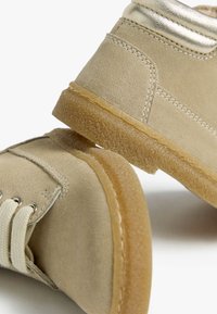 Beige Velourslederschuhe mit strukturierter Oberfläche, ausgestattet mit einer Gummisohle und hellen Schnürsenkeln sowie metallischen Akzenten nahe dem Knöchel.