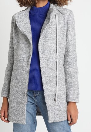 Kvinde iført en lysegrå uldfrakke med lynlås over en blå ribbet rullekravetrøje og blå jeans, stående mod en hvid baggrund.