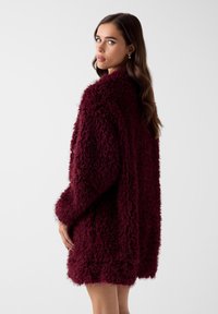 Bordeauxroter Oversize-Kunstpelzmantel mit strukturierter Oberfläche, langen Ärmeln und hohem Kragen. Das Design ist weich und gemütlich.