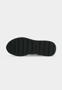 Semelle de chaussure en caoutchouc noir avec des rainures horizontales et le logo « BOSS » embossé au centre sur un fond gris clair.