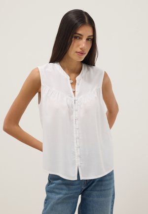 Vrouw met lang donker haar, gekleed in een mouwloze witte blouse met knopen en blauwe spijkerbroek, staand met handen achter haar rug tegen een egale achtergrond.