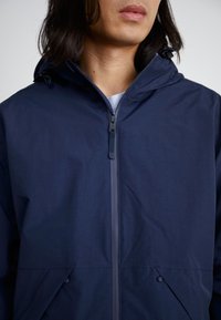 Veste légère bleu marine avec capuche, fermeture éclair frontale et poches latérales. Texture lisse, fermeture à glissière intégrale et cordon de serrage réglable.