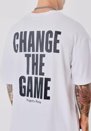 Witte T-shirt met een losse fit, voorzien van grote zwarte tekst op de achterkant: "CHANGE THE GAME" en "Project x Paris" daaronder. Korte mouwen.