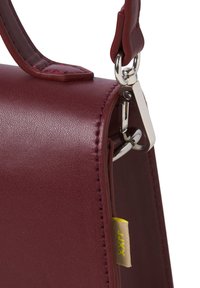 Sac à main en cuir bordeaux avec une forme structurée, agrémenté de coutures contrastées et d'un clip en métal argenté pour l'attache. Détail d'étiquette jaune visible.