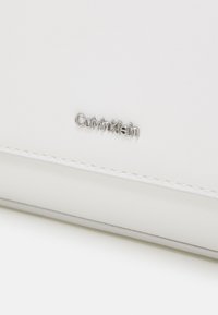 Surface en cuir blanc avec bord cousu et petit logo argenté Calvin Klein centré au-dessus de la couture.