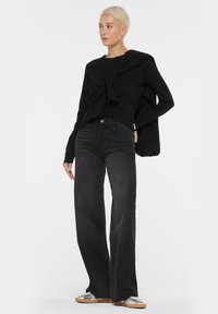 Schwarzes Langarmoberteil mit vorderem Knoten-Detail, kombiniert mit hochgeschnittenen schwarzen Weitbeinjeans und silbernen Sneakers. Einfaches, lässiges Design.