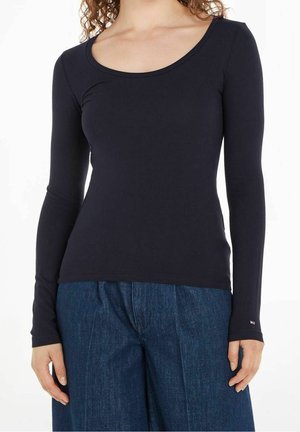 Frau trägt ein eng anliegendes schwarzes Langarmoberteil und lockere blaue Jeanshosen, steht vor einem weißen Hintergrund.