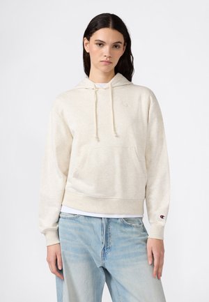 Mujer joven con cabello largo y oscuro, vestida con una sudadera con capucha beige claro y vaqueros holgados azul claro, de pie contra un fondo blanco liso.