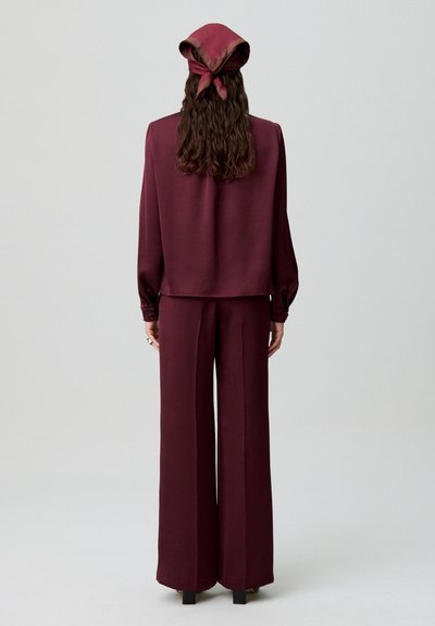 Completo bordeaux abbinato composto da una blusa ampia con maniche a sbuffo e pantaloni a gamba larga, completato da una sciarpa di raso annodata sul retro.