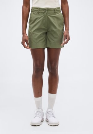 Osoba oblečená v olivovo zelených prispôsobených šortkách, biele ponožky a biele atletické topánky, stojaca na jednoduchom svetlom pozadí.