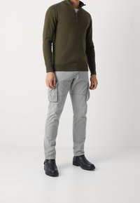 Olivgrüner Zip-Neck-Pullover, hellgraue Cargohose mit Seitentaschen und schwarze Stiefeletten mit strukturierter Sohle, stehend vor einem einfarbigen Hintergrund.