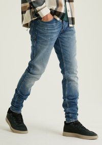Világoskék farmernadrág, slim fit szabásban, enyhe kopással a térdeknél, fekete és zöld sportcipővel párosítva, amelynek mintás talpa van.