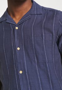 Camicia button-up blu realizzata in tessuto testurizzato con righe verticali bianche, dotata di tasca a toppa e colletto da campeggio.