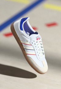 Baskets blanches de sport avec des rayures bleu et rouge, présentant un capuchon en daim, une semelle en caoutchouc et un col rembourré avec une étiquette de logo.