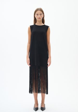 Schwarzes ärmelloses Kleid mit geradem Schnitt und texturiertem Fransenabschluss. Glattes Material und einfacher Ausschnitt. Kombiniert mit schwarzen spitz zulaufenden Absätzen.