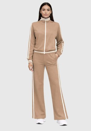 Femme portant une veste de sport beige à fermeture éclair assortie à un pantalon large avec des bandes blanches et des baskets blanches, debout devant un fond gris.