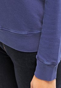 Paarse katoenen sweatshirt met een geribde hals en manchetten, met een gladde textuur en gestikte zomen. Draagt met een donkere spijkerbroek.