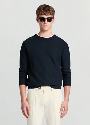 Maglione blu navy a costine con maniche lunghe e scollo rotondo, indossato sopra una camicia bianca, abbinato a pantaloni sartoriali color crema e occhiali da sole scuri.