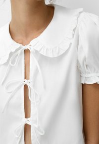Vit blus med en volangkrage, korta puffärmar och knytning fram i ett mjukt tyg. Hög hals med öppen nyckelhål-detalj.