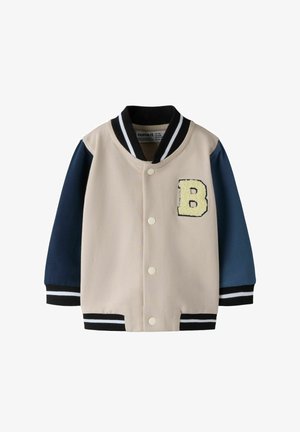 Børns beige og marineblå varsityjakke med sorte og hvide stribede manchetter og krave, trykknapper og et gult bogstav B-patch på brystet.