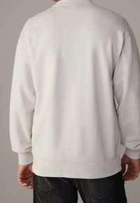 Sudadera blanca con cuello alto, puños acanalados y un corte relajado. Textura suave y diseño minimalista, combinada con jeans oscuros.