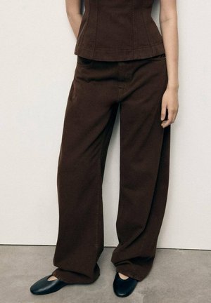 Žena nosící tmavě hnědé kalhoty s širokými nohavicemi a odpovídající tílko bez rukávů s černými nízkými botami, stojící před jednoduchou světlou stěnou.