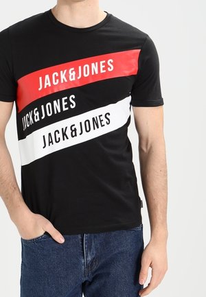 Mann trägt ein schwarzes T-Shirt mit drei diagonalen Streifen in Rot, Schwarz und Weiß, jeweils mit dem Schriftzug "JACK & JONES", kombiniert mit blauen Jeans.