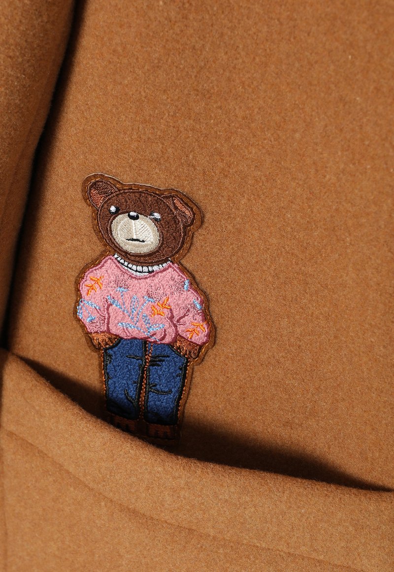 Brązowy materiał z haftem niedźwiedzia w różowym sweterku i niebieskich dżinsach, z kwiatowymi detalami i okrągłym dekoltem.