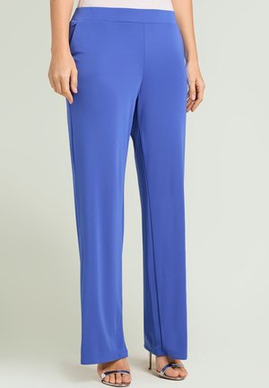 Kobieta nosząca proste, królewskie niebieskie spodnie w połączeniu ze srebrnymi sandałami na wysokim obcasie z odkrytymi palcami, stojąca na jednolitym jasnym tle.