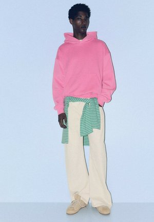 Jeune personne debout portant un sweat à capuche rose vif, un pantalon large beige, des chaussures beiges et un pull vert rayé noué autour de la taille sur un fond uni.