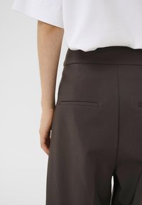 Pantaloni marroni su misura con una texture liscia, dotati di due tasche posteriori. Indossati con una blusa bianca oversize, parzialmente visibile nell'immagine.