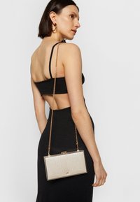 Femme portant une robe noire dos nu et des boucles d'oreilles créoles dorées, tenant une petite pochette métallique texturée rectangulaire avec une bandoulière chaîne dorée.