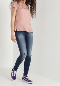 Top de manga corta rosa claro, jeans ajustados azules con detalles desgastados y zapatillas moradas con cordones blancos. Conjunto casual presentado.