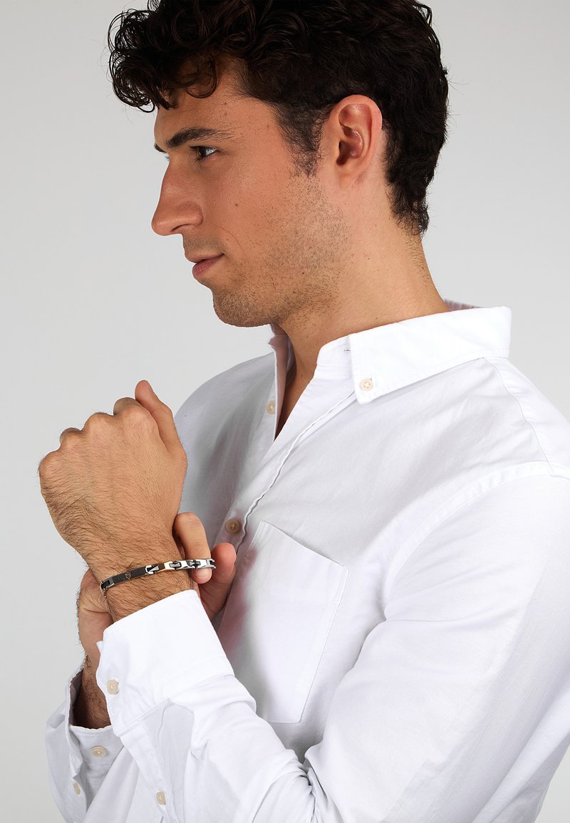 Camicia bianca con bottoni e tasca sul petto, indossata da un modello maschile. Un braccialetto a catena nero e argento adorna il suo polso, evidenziando un design resistente.