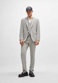 Light grey tailored suit with a textured finish, white shirt underneath, and a black cap with "HUGO" in red lettering. Black chunky shoes.